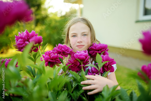 Cute young girl admiring tall purple peonies on sunny summer day. Child and flowers, summer, nature and fun.
