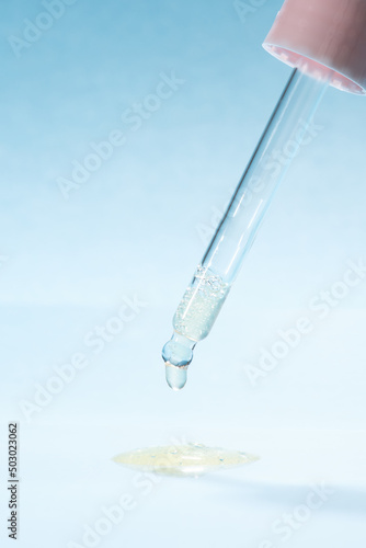 Transparent pink pipette dropper with light golden drop of serum cosmetics, oil, gel ou cream on blue  background and bubbles of air. Minimal beauty concept, macro closeup, front view