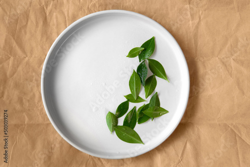 Top down view mockup empty ceramic white color round plate with water decoration setting with fresh tropical green leaves on creased brown paper background to be use for skincare or food products