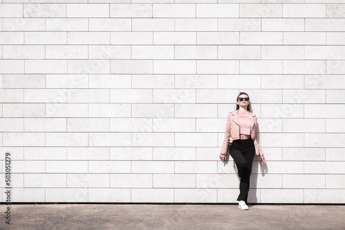 Beauty, lifestyle and fashion concept. Beautiful woman with pink blouse and jacket, black pants, white sneakers and sunglasses standing in front of huge brick wall background with copy space.
