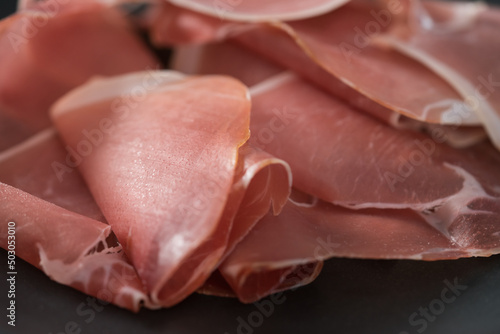 Thin italian prosciutto slices on black plate on conctere background closeup