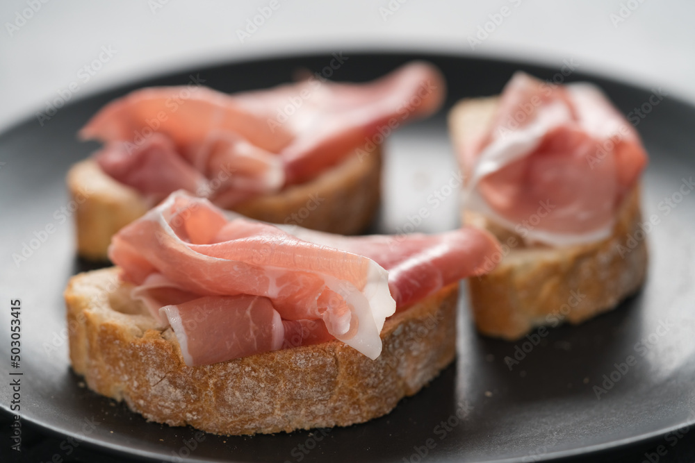 ciabatta with thin prosciutto on black plate on conctere background