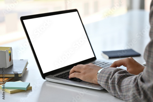 Close up side view of an adult female in a suit typing something with an empty monitor screen of a laptop on a busy white desk. Business and Technology concepts.