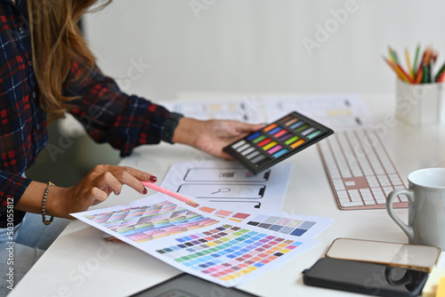 The close-up of a female designer choosing a color tone for the new media development projects with stationery and office suppliers on a desk. Technology and design concepts.