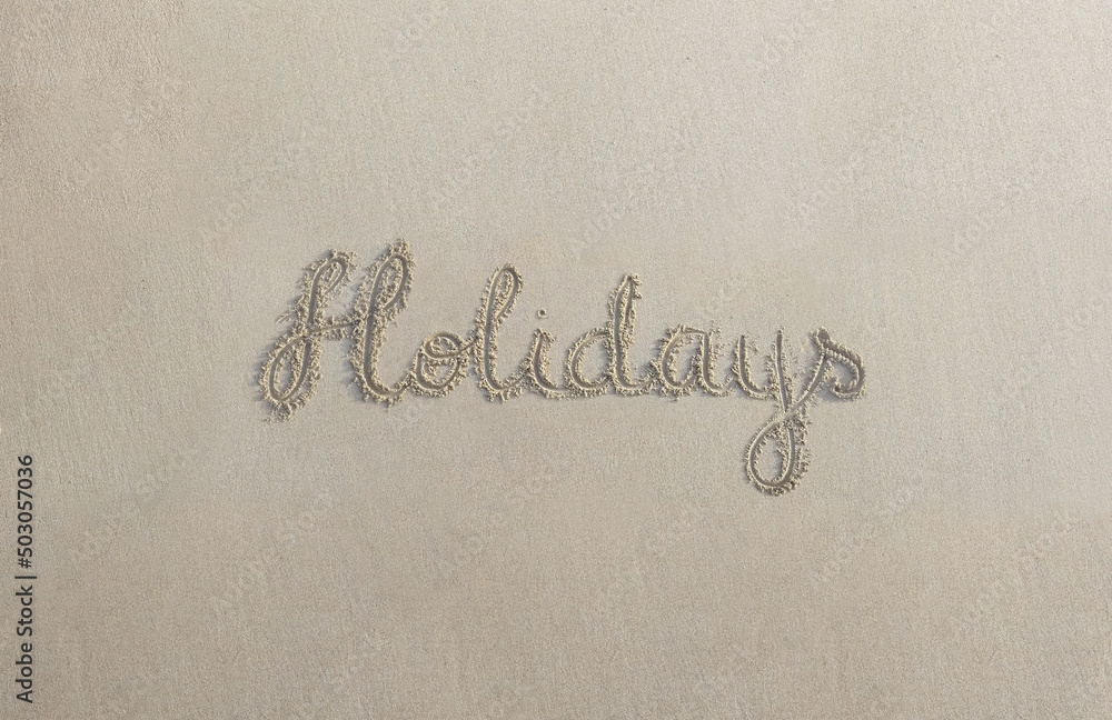 “Holidays” handwritten on sand at the beach on a warm summer day “Holidays” handwritten on sand at the beach on a warm summer day