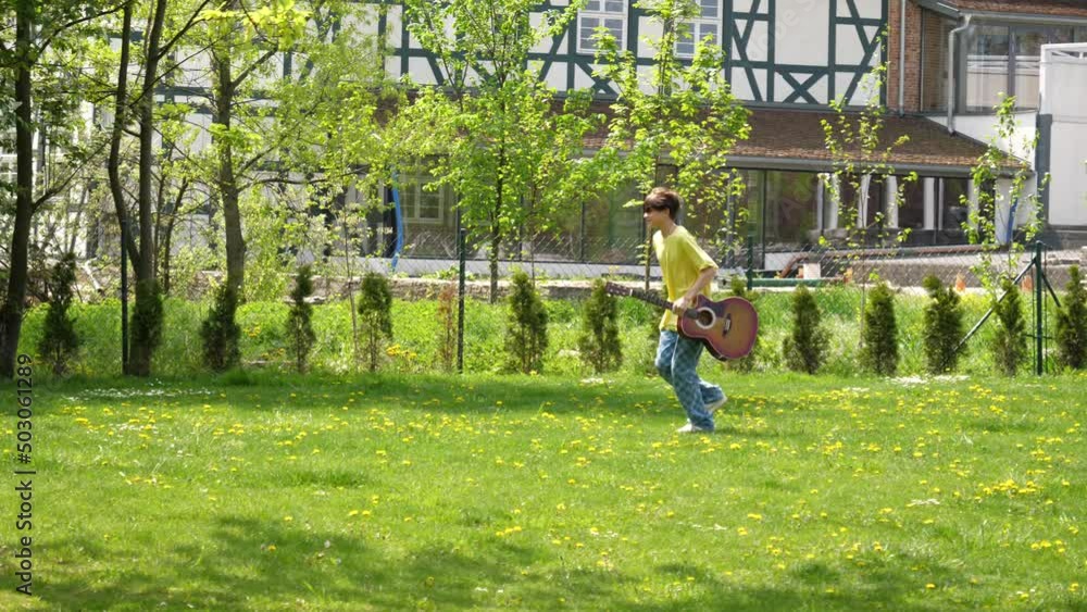 Teenager boy with guitar run through green park lawn hurrying somewhere Teenager boy with guitar run through green park lawn hurrying somewhere