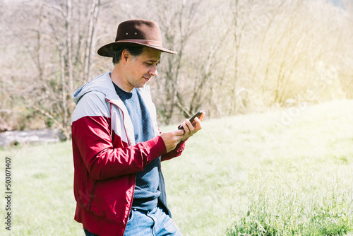 Entrepreneurial self-employed man wearing a hat, looking at his mobile phone while relaxing in the field, on a meadow. Concept of work, enjoy, relax, Internet, technology and connectivity.