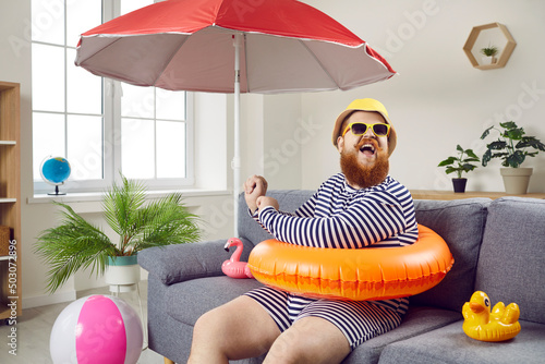 Summer party at home. Funny chubby man having fun sitting on sofa in living room with inflatable swimming circle. Humorous man has absurd vacation under beach umbrella on improvised home beach. photo