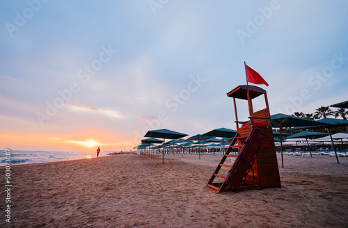 Fototapeta Naklejka Na Ścianę i Meble -  Red riscue flag on lifeguard tower on sea beach at sunset time.