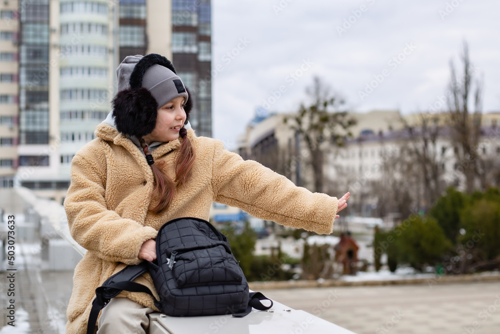a cute seven-year-old girl with a backpack in her hands is sitting on the parapet in the city after school a cute seven-year-old girl with a backpack in her hands is sitting on the parapet in the city after school