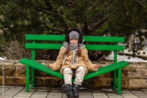 a cute six-year-old girl in a beige fur coat made of eco-friendly faux fur is sitting on a bench