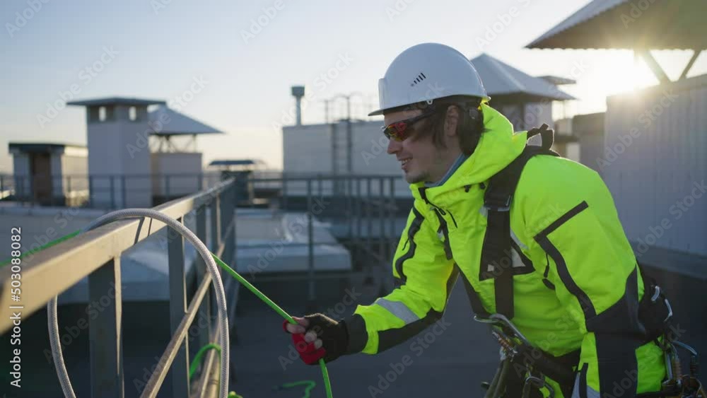 Smiling climber finishes work and gathers equipment at sunset. Industrial climber removes special green rope from building after work closeup Smiling climber finishes work and gathers equipment at sunset. Industrial climber removes special green rope from building after work closeup