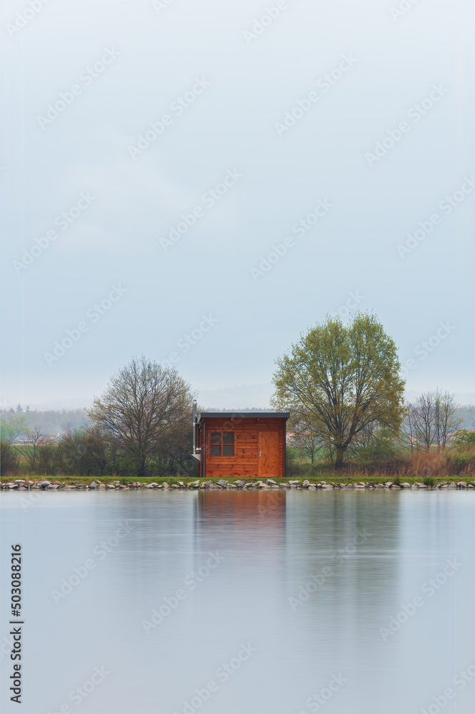 Obraz premium Wooden cabin, building on pond shore in rain moody weather. Long exposure, Czech landscape