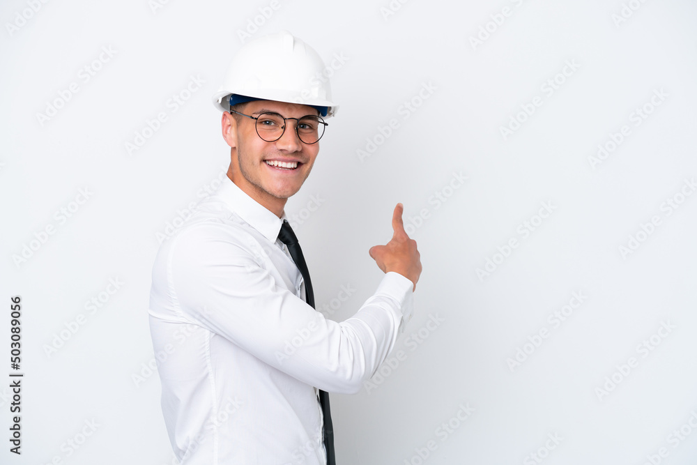 Young architect caucasian man with helmet and holding blueprints isolated on white background pointing back