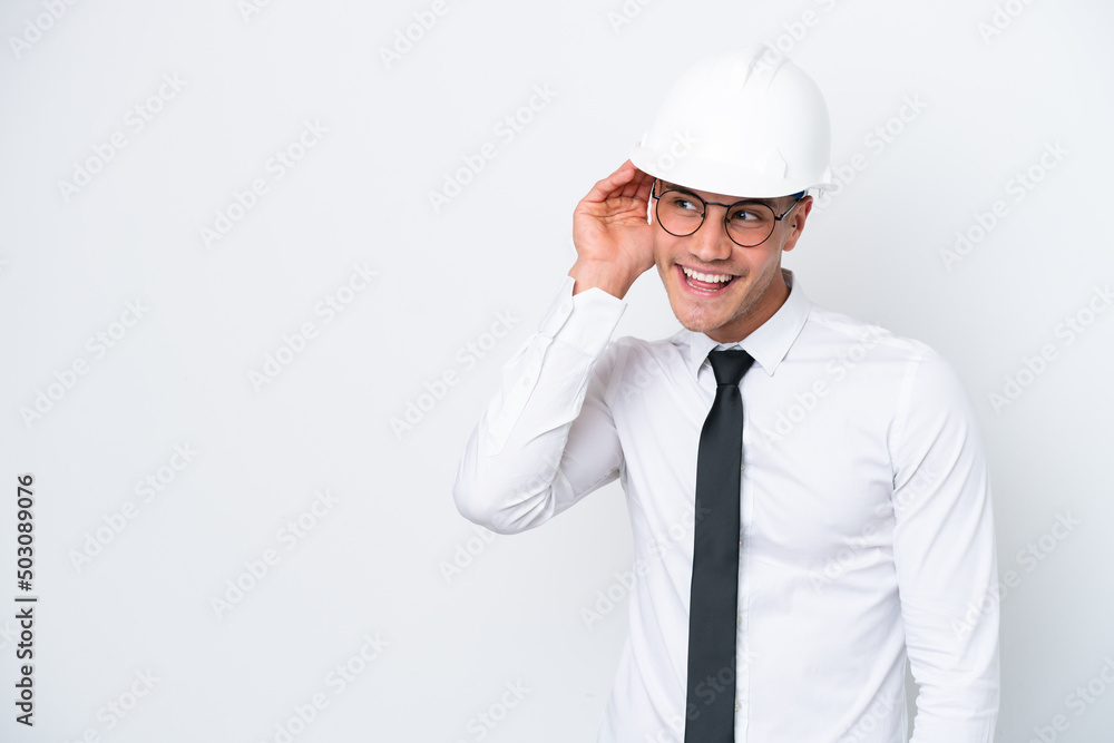 Young architect caucasian man with helmet and holding blueprints isolated on white background listening to something by putting hand on the ear