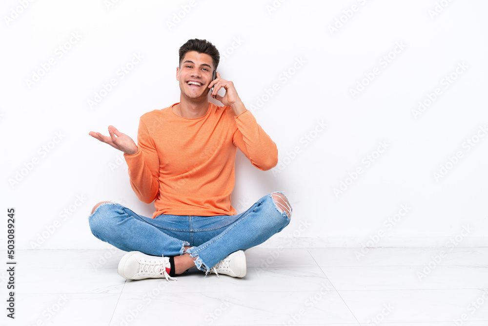 Young man sitting on the floor isolated on white background keeping a conversation with the mobile phone with someone Young man sitting on the floor isolated on white background keeping a conversation with the mobile phone with someone