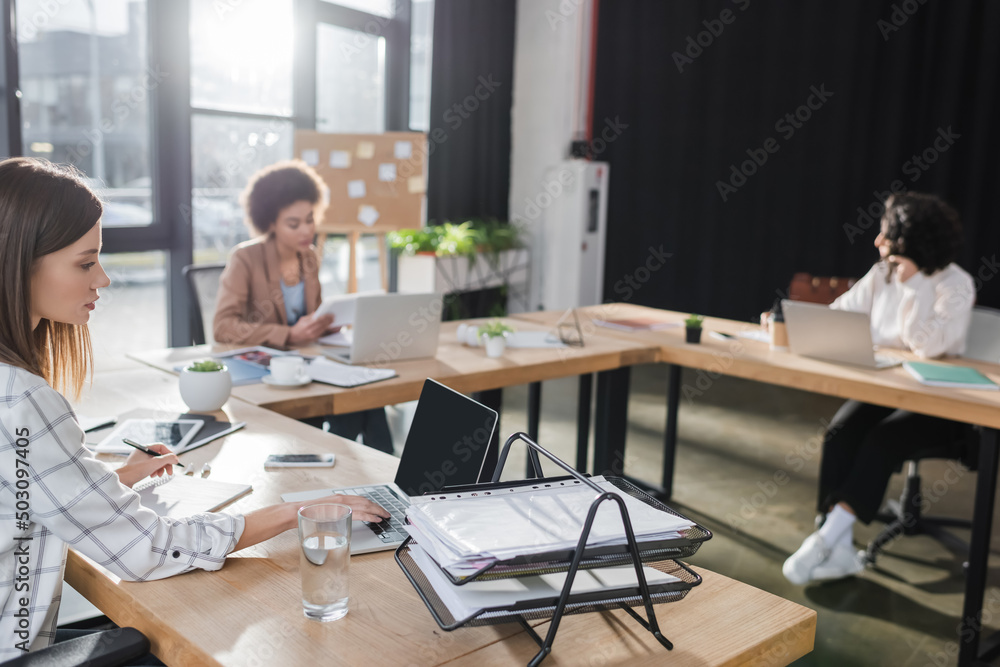 Young businesswoman using laptop near documents and water in office.