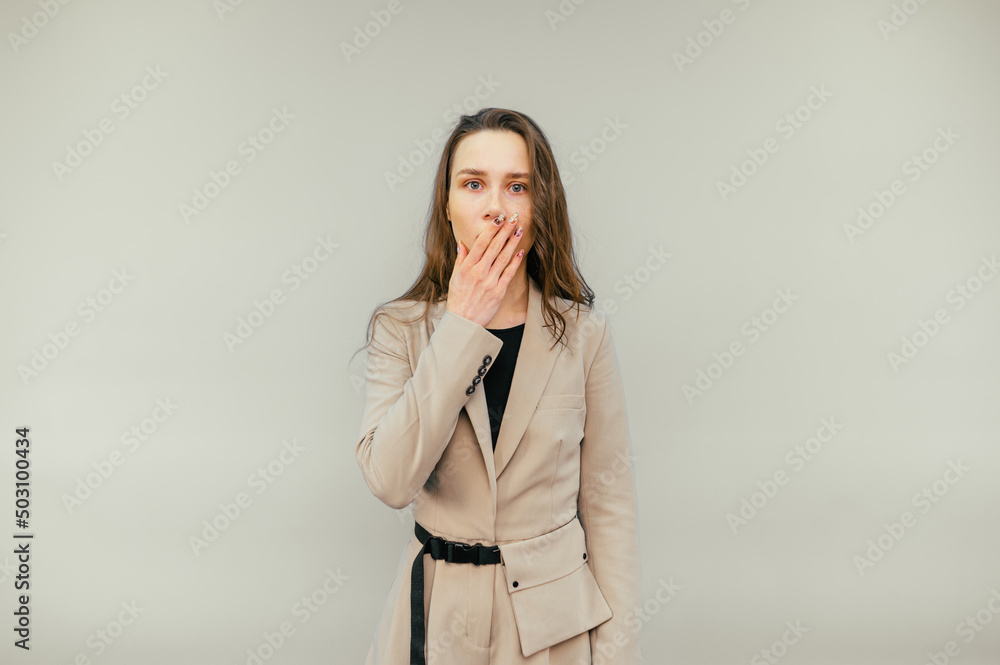Shocked woman in a suit stands on a beige background and covers her face with her hand and looks at the camera with a surprised face. Shocked woman in a suit stands on a beige background and covers her face with her hand and looks at the camera with a surprised face.