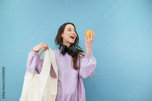 Attractive woman with an eco bag in her hands stands on a blue background with an apple in her hand and looks at the fruit with a smile on her face.