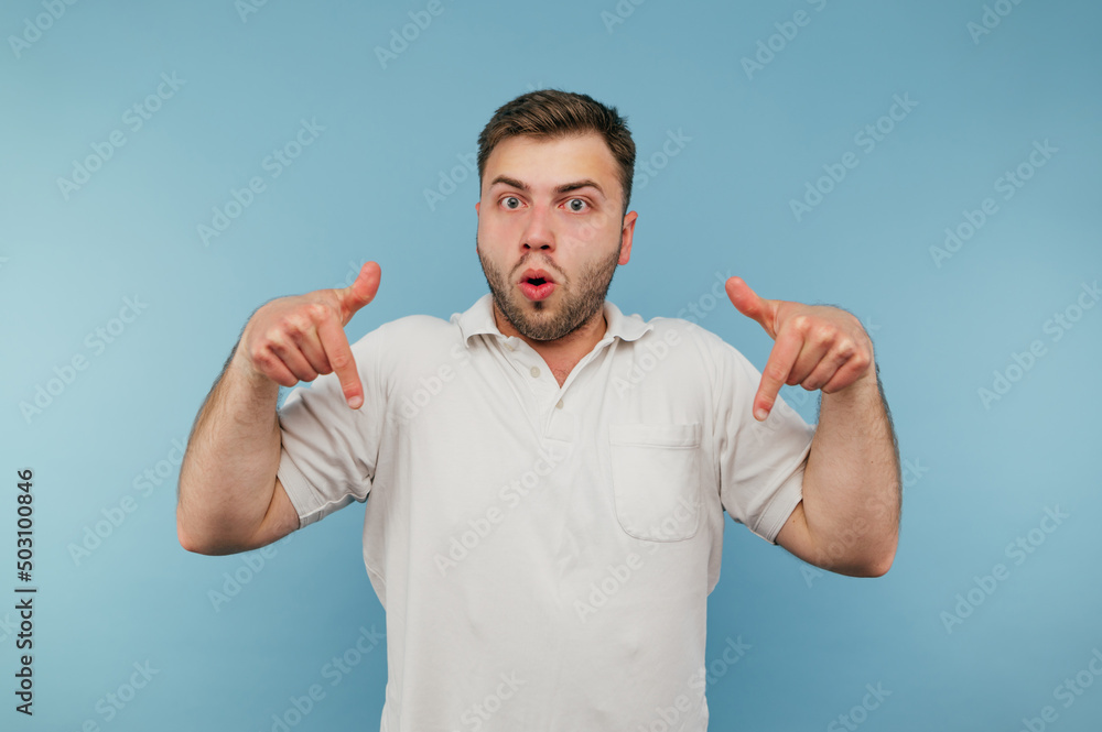 Surprised man with bristles and in a white T-shirt stands on a blue background and shows his fingers down on an empty spot and looks at the camera with a shocked face Surprised man with bristles and in a white T-shirt stands on a blue background and shows his fingers down on an empty spot and looks at the camera with a shocked face
