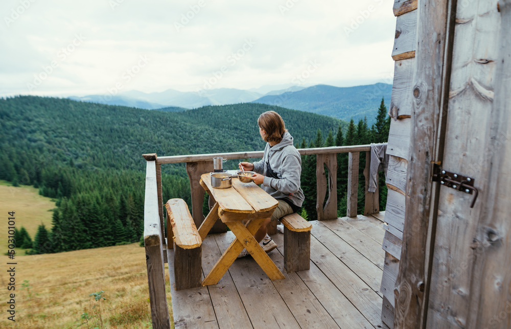 The tourist guy sits at a table on the terrace of a country wooden house and eats against the backdrop of mountain scenery.