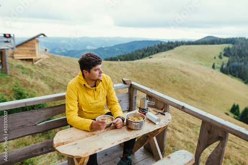 Male tourist sitting at a table on a terrace in the mountains and eating pasta cooked on a burner.