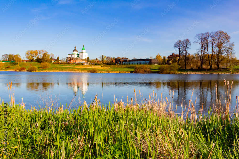 Any pond with clear, blue water on a bright, sunny day in an agricultural area and with a reflection in the water of a white, Orthodox church standing on a hill.