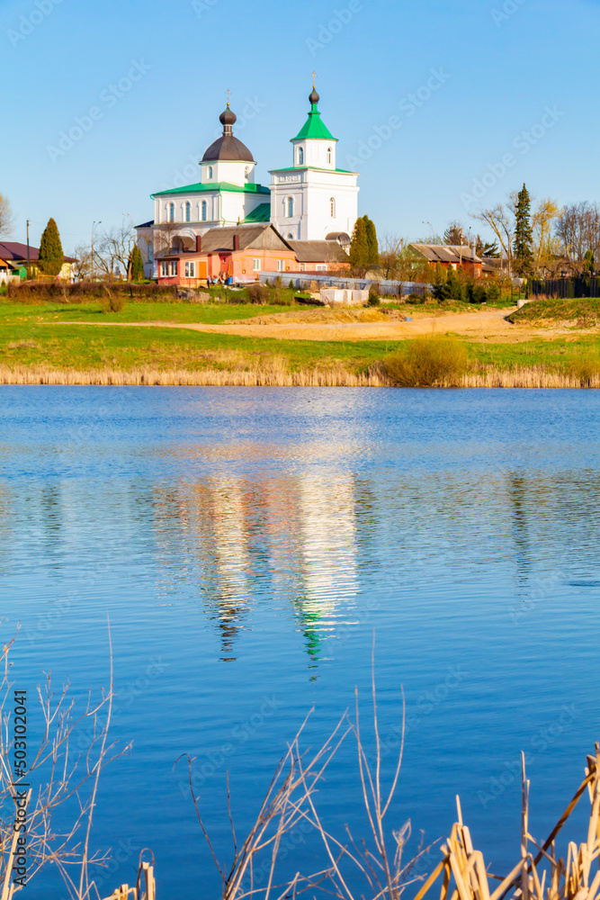 Any pond with clear, blue water on a bright, sunny day in an agricultural area and with a reflection in the water of a white, Orthodox church standing on a hill.