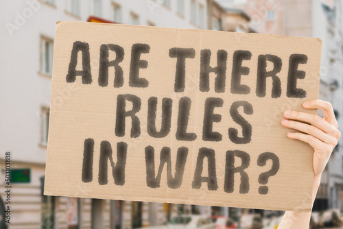 The question " Are there rules in war? " is on a banner in men's hands with blurred background. Evil. Failure. Injury. Break. Offense. Criminality. Dishonesty. Violation. Sin. Illegality. Fighting