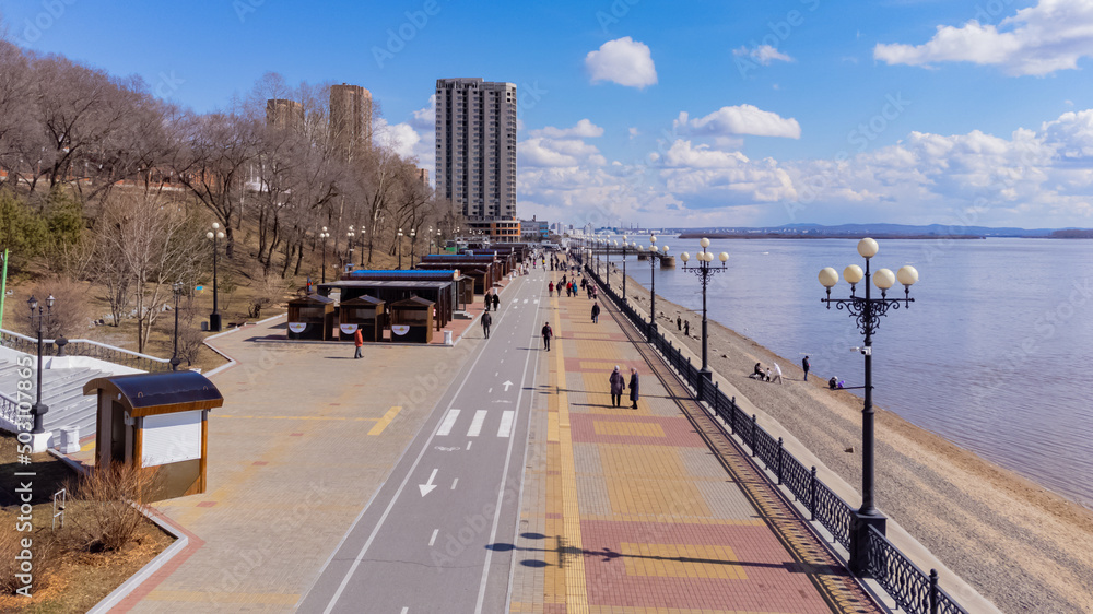 City embankment on the Amur River in Khabarovsk in early spring. A sunny day. The ice on the river has already melted. The trees haven't turned green yet. People are walking along the embankment. 