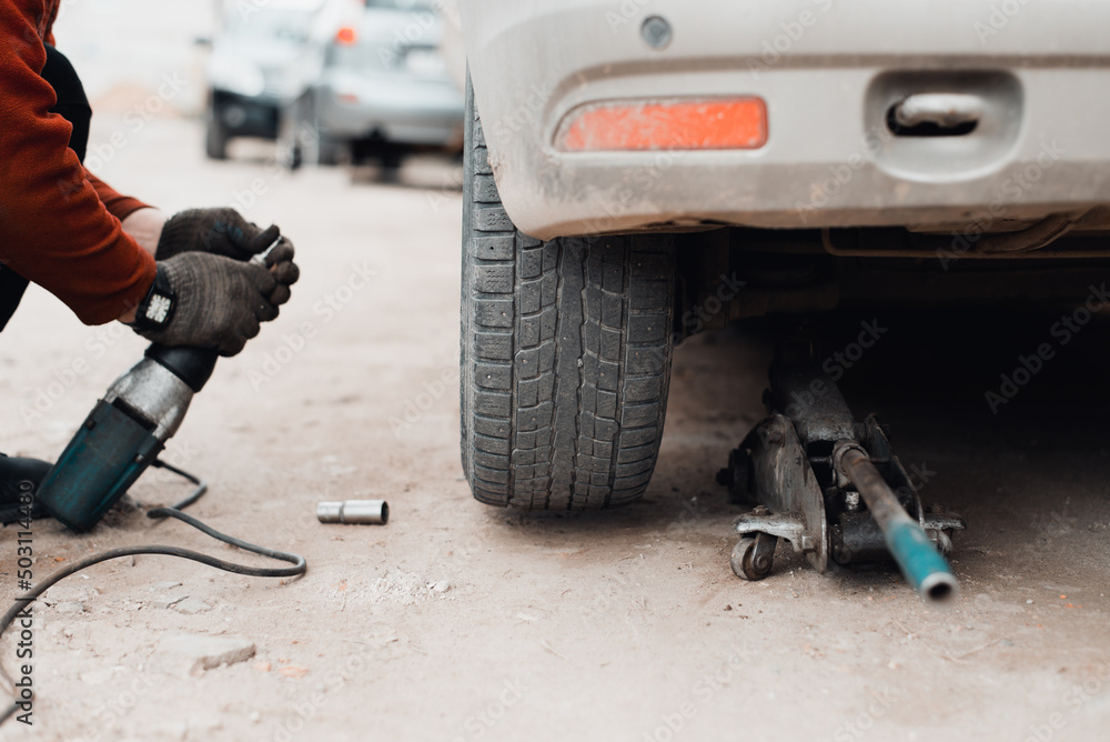 Mechanic using electric wrench repairing car on jack outdoors, low angle view. Broken vehicle repair, problem or wheel replacement concept. Selective focus on wheel