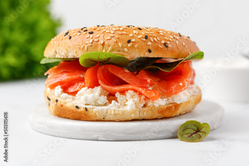 Bagel sandwich with salmon, cream cheese and green leaves on marble board and white background. Burger with salmon