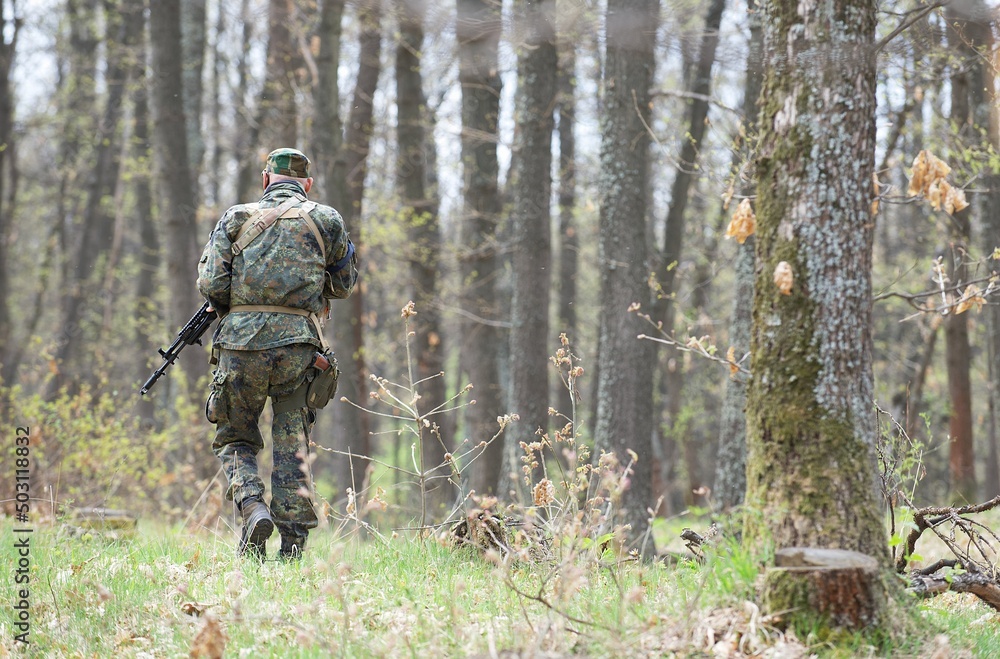 The figure of a man from the back walking in a camouflage green uniform with ammunition with weapons for the game air soft moves towards the forest between the trees in spring forest.