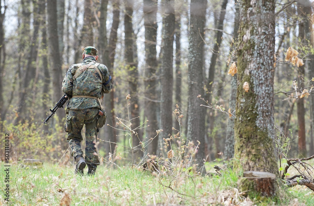 The figure of a man from the back walking in a camouflage green uniform with ammunition with weapons for the game air soft moves towards the forest between the trees in spring forest. The figure of a man from the back walking in a camouflage green uniform with ammunition with weapons for the game air soft moves towards the forest between the trees in spring forest.
