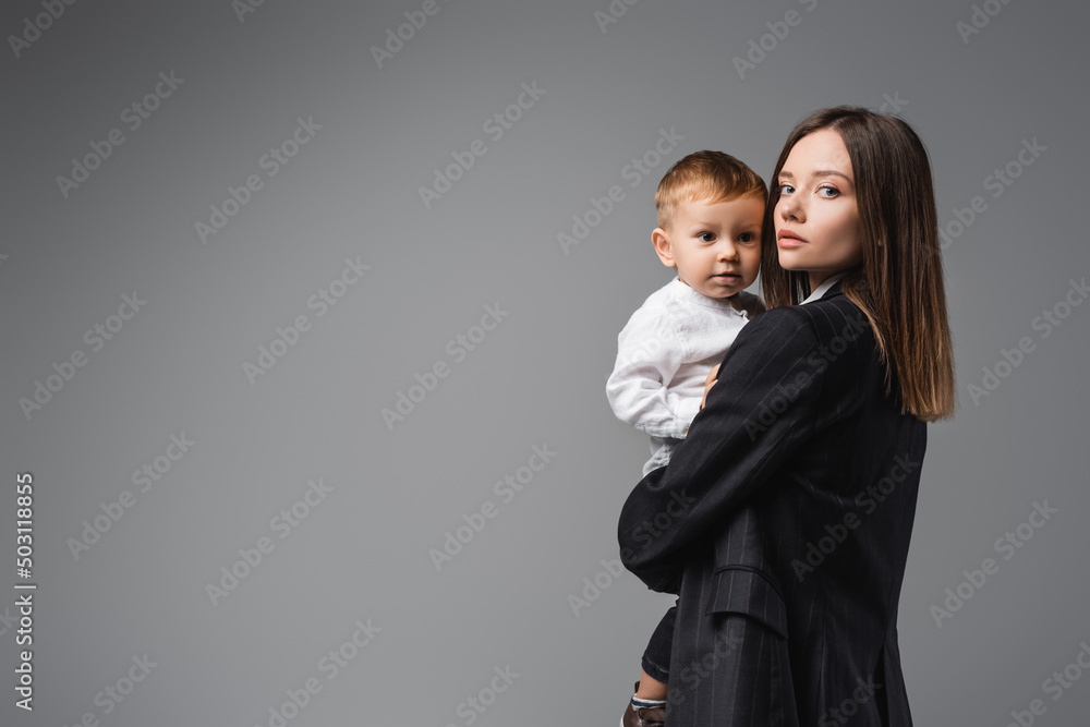 woman in black blazer holding toddler son and looking at camera on grey.