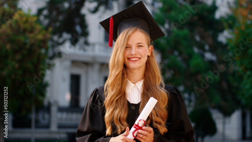 Beautiful with a large smile lady graduate posing in front of the camera while holding her diploma in the college garden