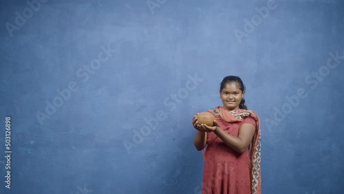 Indian poor girl saving money by placing coins on piggy bank or pot gullak by looking at camera - concept of financial, future education and banking. photo