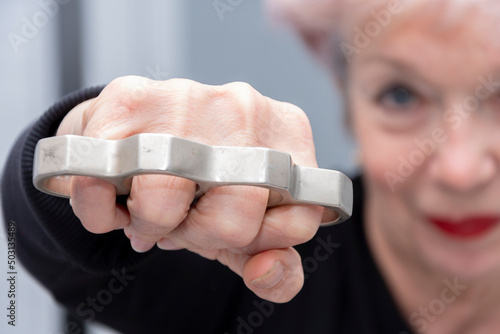 An elderly woman strikes with brass knuckles on a light background, close-up, selective focus. Concept: domestic brawl, family scandal, quarrel with her husband, grievous bodily harm.