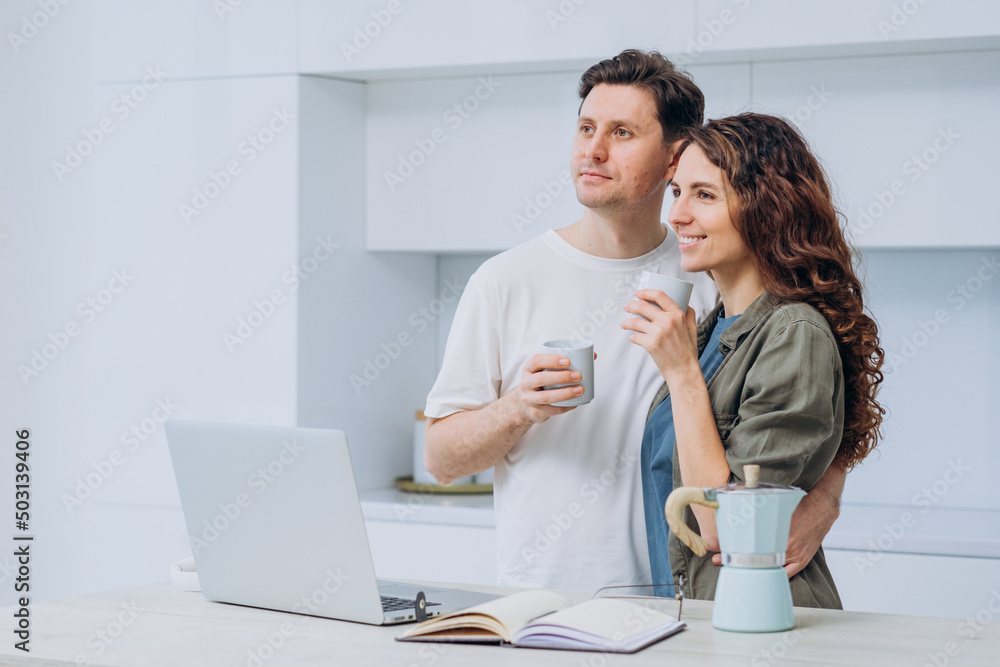 Married couple of freelancers took a coffee break in a bright kitchen standing at a table with a laptop. Happy man and woman stand embracing and drinking aromatic coffee. Married couple of freelancers took a coffee break in a bright kitchen standing at a table with a laptop. Happy man and woman stand embracing and drinking aromatic coffee.