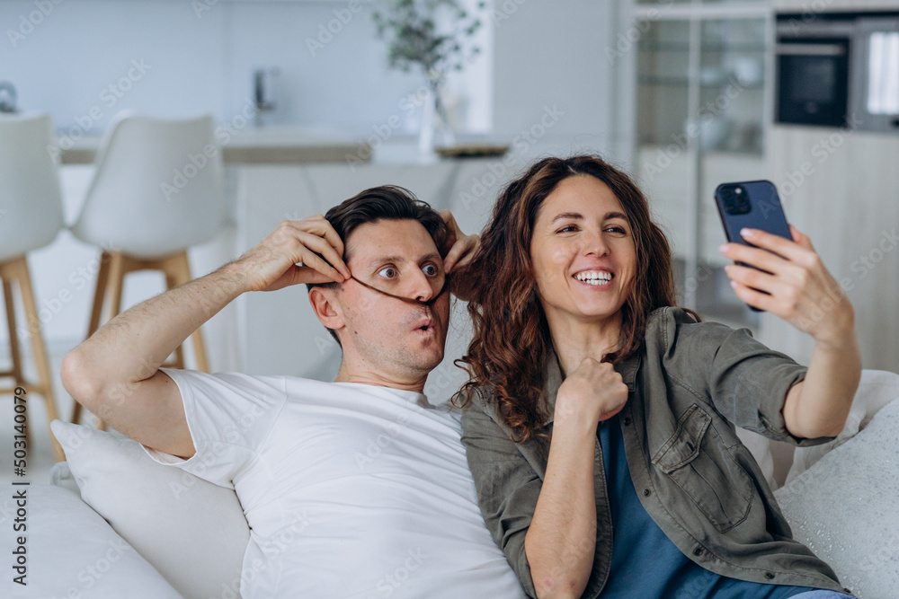 Cheerful in love couple taking a selfie on the phone while sitting on the couch together, making funny mustaches out of their hair and taking pictures Cheerful in love couple taking a selfie on the phone while sitting on the couch together, making funny mustaches out of their hair and taking pictures