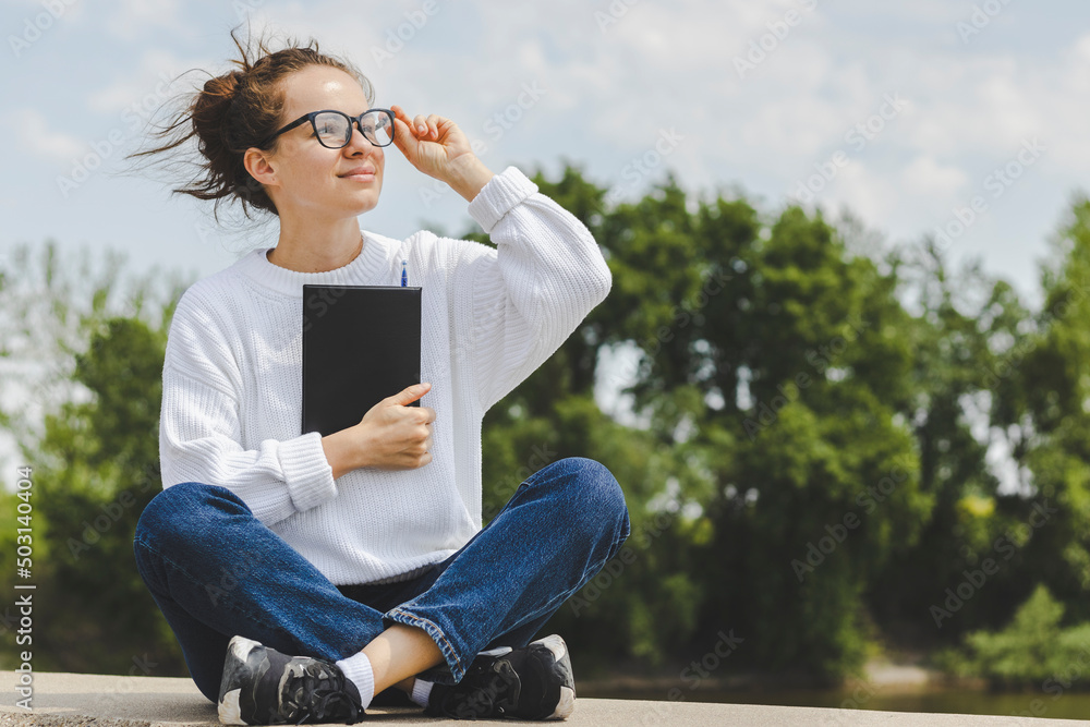 Young adult female student in glasses with notebook in her hands outdoors.