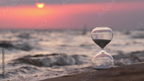 Sand-glass stand on sand against small waves of sea. Selective focus on foreground, blurred background. Continuous motion of waves, slow flow of sand grains in hourglass. Sun moves down and appears photo