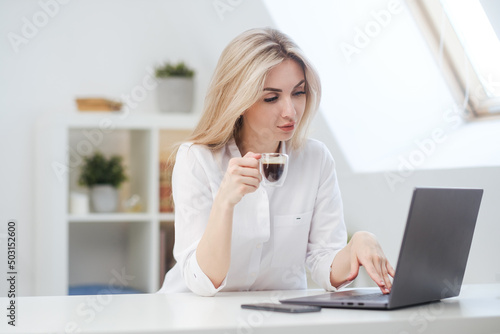 Young beautiful caucasian blonde woman working on laptop at home and drinking coffee. Remote work or freelance. © spaskov