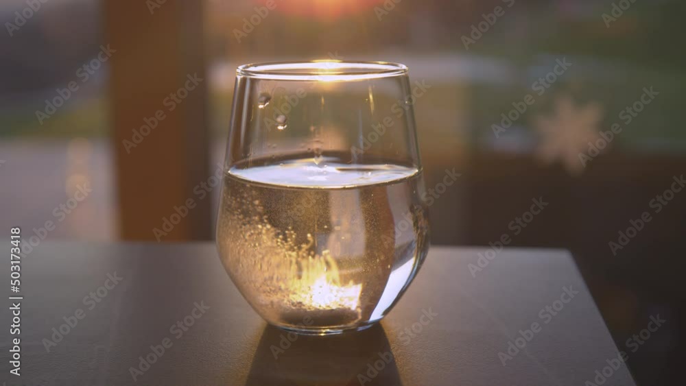 SLOW MOTION, CLOSE UP: Dissolving effervescent tablet dropped in a drinking glass backlit with the golden setting sun. Sparkling beverage for regaining health. Home treatment for seasonal illnesses.