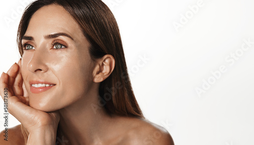 Smiling female aging model, clear glowing skin, nourished face without blemishes, applies daily cream, hyaluron anti-aging treatment, looking in mirror at herself, white background