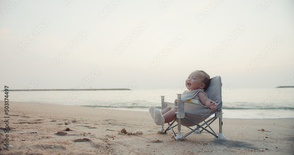 happy cute adorable Asian baby infant sitting relaxing on little chair and smiling with waves on background at seaside tropical sandy beach in sunrise during holiday vacation summertime Thailand