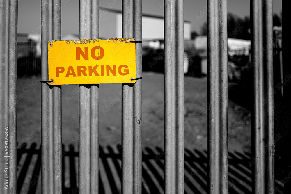 Shallow focus of a small No Parking sign seen on a metal gated entrance to an industrial yard. The sign is showing weathering around the edges. Shallow focus of a small No Parking sign seen on a metal gated entrance to an industrial yard. The sign is showing weathering around the edges.