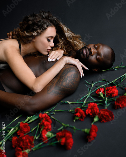 Cropped of passionate woman with Afro hairstyle, wearing in bra, lying on naked handsome tattooed Afro American man with closing eyes near scattered red carnations photo