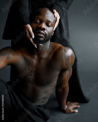 Cropped of calm Afro American male model with closed eyes and tattoos on muscular arms, sitting on floor in isolated studio while hands of faceless woman with white manicure touching his face