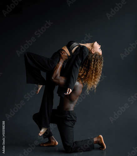 Side view of strong dark-skinned man in black trousers, leaning on knee while lifting sensual woman in black costume and laced lingerie while having rehearsing of dance against black backround photo